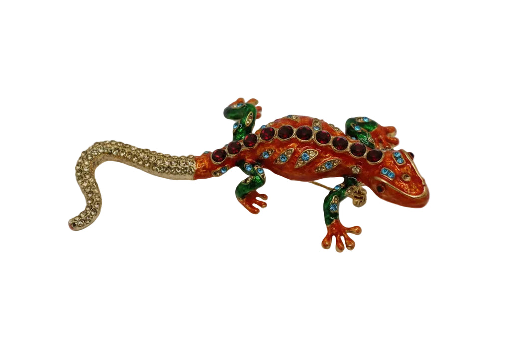 A decorative lizard-shaped brooch featuring a gold-toned textured tail, green and orange body with blue and red rhinestone accents, and small red rhinestones along the back, displayed on a neutral light gray background.